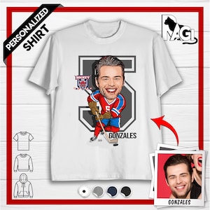 May include: Personalized white t-shirt with a cartoon illustration of a hockey player wearing a red and blue jersey with the number 5. The player is holding a hockey stick and is standing in front of a gray number 5. The text "GONZALES" is printed below the player. The t-shirt is available in white, gray, navy, and black.