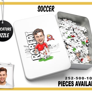 May include: A white metal tin with a caricature of a soccer player on the front. The tin contains a jigsaw puzzle with 252, 500, or 1000 pieces. The text "SOCCER" is at the top of the tin and "CARICATURE PUZZLE" is on a black circle at the top left of the image. The text "ROBERT" is written vertically on the left side of the caricature. The text "252-500-1000 PIECES AVAILABLE" is at the bottom of the tin.