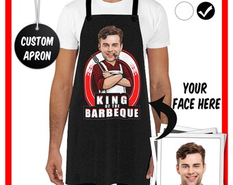 May include: Black apron with a cartoon illustration of a person holding a fork and a spatula. The text "King of the Barbeque" is printed on the apron. A photo frame with a space for a photo is on the right side of the image.