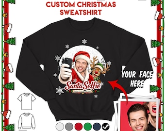 May include: A black long-sleeve sweatshirt featuring a graphic of a person in a Santa hat taking a selfie with a reindeer. The text reads "CUSTOM CHRISTMAS SWEATSHIRT" at the top and "Santa Selfie" below the graphic, with "YOUR FACE HERE" pointing to a photo.