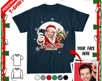 May include: A navy blue short-sleeve t-shirt featuring a graphic of a person in a Santa hat taking a selfie with a reindeer. The text reads Santa Selfie Making Christmas Great Again. The design includes white snowflakes on a dark blue circular background.
