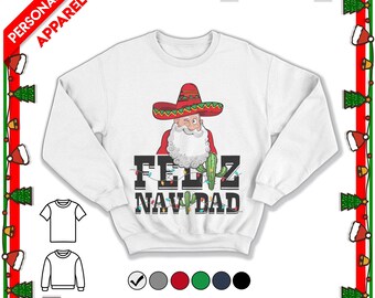 May include: A white long-sleeve sweatshirt featuring a graphic of Santa Claus wearing a red and green sombrero. Below the graphic, the text reads FELIZ NAVDAD in black block letters, with a green cactus and colorful Christmas light strings.