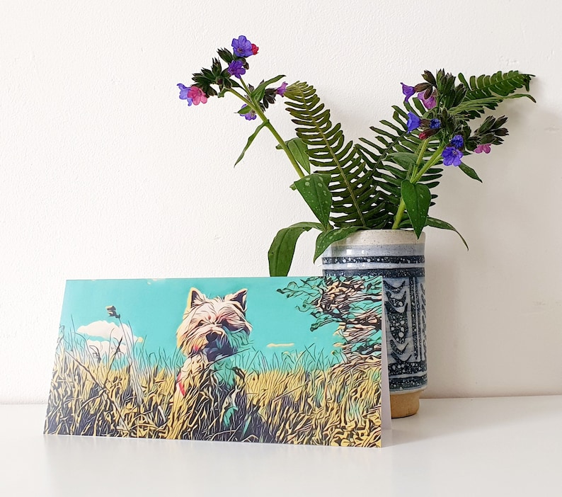May include: A greeting card featuring a cartoon illustration of a white dog with brown ears in a field of green grass. The dog is looking to the right. The background is a light blue sky with white clouds. The card is blank inside.