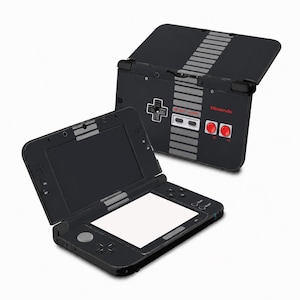 May include: A black handheld gaming console featuring a design inspired by a classic controller, including a grey striped pattern, a black directional pad, and two red circular buttons. The console is shown both open and closed, with the Nintendo logo.
