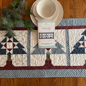 May include: A quilted table runner with a blue and white gingham pattern and three red, white, and blue patchwork pine trees. The runner is 15" x 48" and is called "Country Pines".