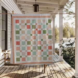 May include: A patchwork quilt with a square pattern in shades of coral, green, and cream, hanging on a washing line. The quilt has a light peach border and is displayed outdoors on a wooden porch.