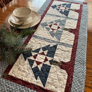 May include: A quilted table runner with a blue and red patchwork design featuring three stylized Christmas trees. The runner has a blue and white gingham border.