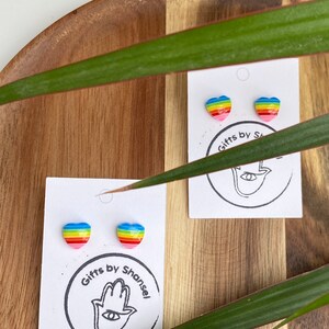 Könnte beinhalten: Zwei Paare Regenbogen-Herz-Ohrringe auf weißen Karten mit dem Text "Gifts by Shansel" und einem Hamsa-Handsymbol.