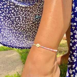 May include: A beaded anklet featuring a small white flower charm and pastel-coloured beads. The anklet is worn on an ankle, with a blue floral dress visible in the background. The anklet is a delicate piece of jewellery.
