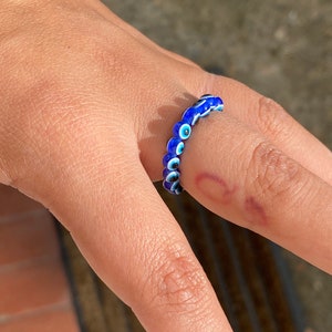 Op de afbeelding: Een blauwe en witte ring met kralen in de vorm van een boze oog. De ring is gemaakt van kleine, ronde kralen met een blauwe achtergrond en een wit oog in het midden.