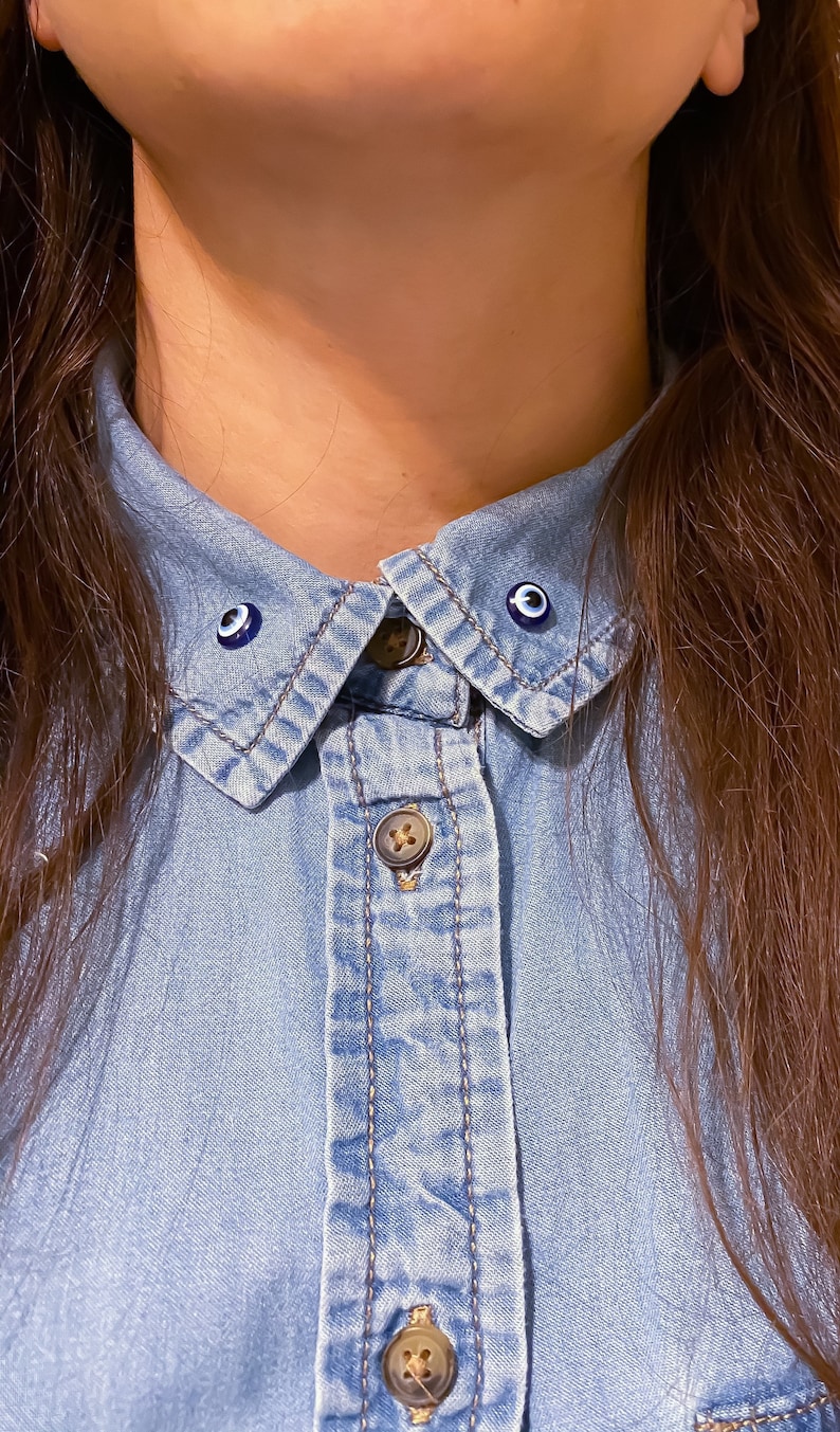 May include: A blue denim shirt with two blue evil eye pins on the collar.