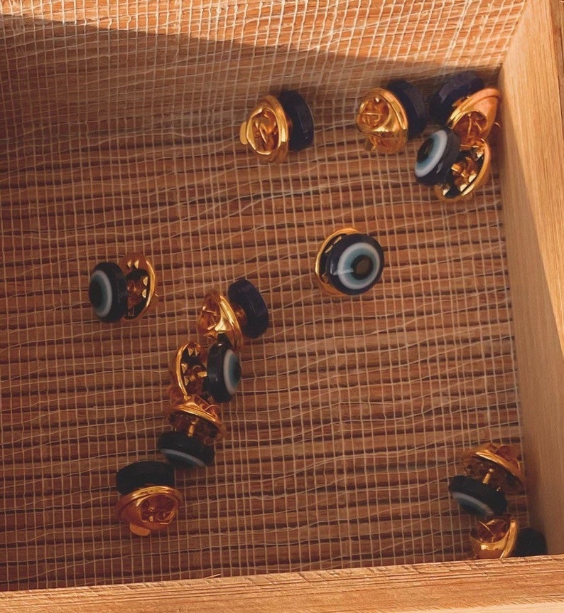 May include: A wooden box filled with small, gold-colored pins with blue and white evil eye charms.