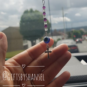 May include: A silver chain with purple and white beads, a blue evil eye charm, and a silver cross charm. The charm is hanging from a rearview mirror.