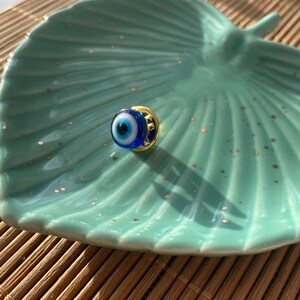 May include: A small blue evil eye charm with a gold setting. The charm is on a light blue and white ceramic dish.