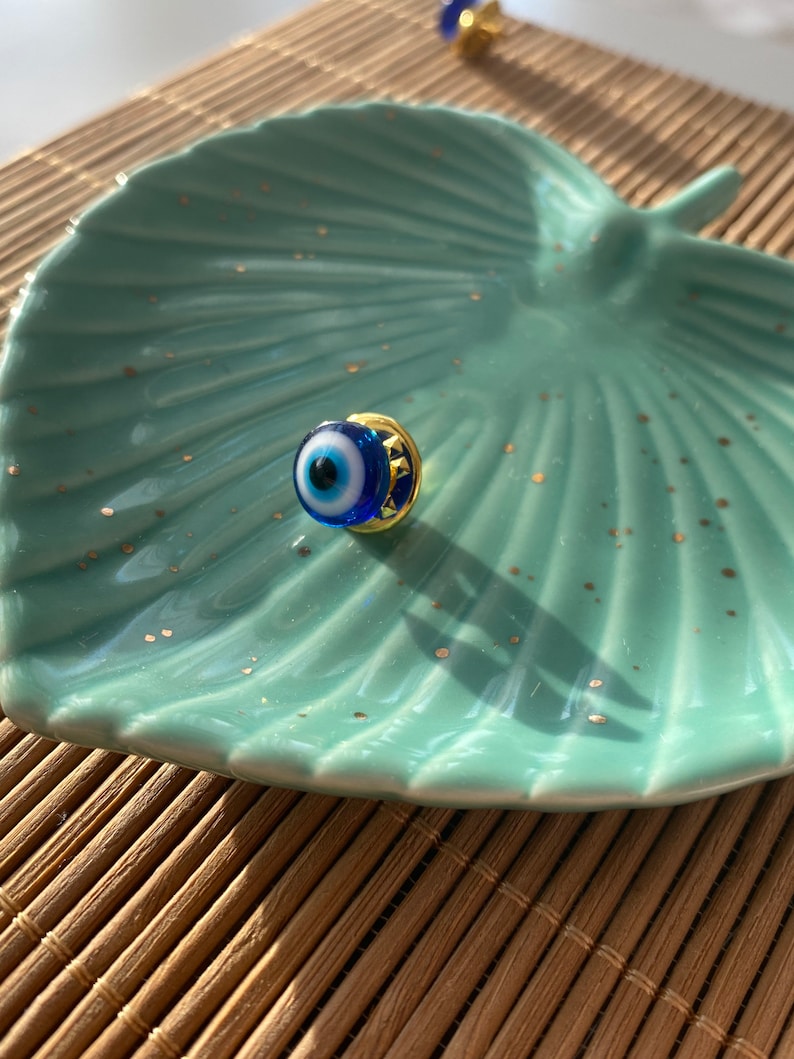 May include: A small blue evil eye charm with a gold setting. The charm is on a light blue and white ceramic dish.