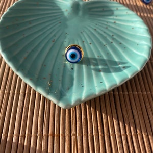 May include: A blue and white evil eye charm with a gold rim on a light blue leaf-shaped dish. The dish is on a woven bamboo mat.