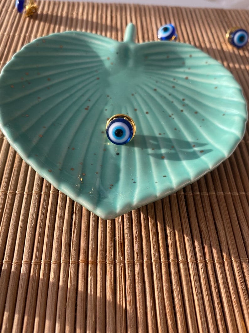 May include: A blue and white evil eye charm with a gold rim on a light blue leaf-shaped dish. The dish is on a woven bamboo mat.