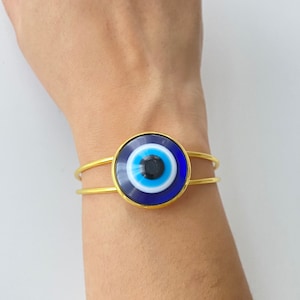Könnte beinhalten: Goldarmband mit einem blauen bösen Blick-Anhänger. Der Anhänger ist rund und hat ein weißes und schwarzes Augen-Design.