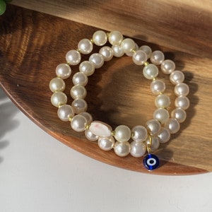 May include: A white pearl bracelet with a gold charm in the shape of a blue evil eye. The bracelet is on a wooden surface.