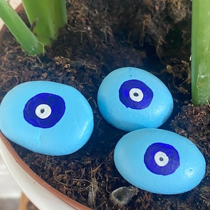 May include: Three light blue painted stones with a dark blue and white eye design. The oval-shaped stones are placed in a white pot with soil and green plant stems.