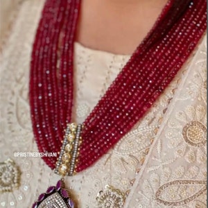 May include: A multi-strand beaded necklace in shades of red, featuring a decorative pendant with a large red gemstone and a silver and gold-toned setting. The necklace is worn over a cream-colored garment with intricate embroidery.
