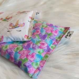 May include: Two fabric eye pillows, one with a white background and a gold unicorn design, and the other with a pink, blue, and green heart pattern. The pillow with the hearts has a label that says "Pretty Eye Pillows".