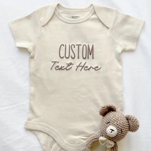 May include: A cream-colored baby onesie with the words "CUSTOM Text Here" in brown script. A small, crocheted teddy bear with a bow tie rests near the onesie. The onesie has short sleeves and snap closures.