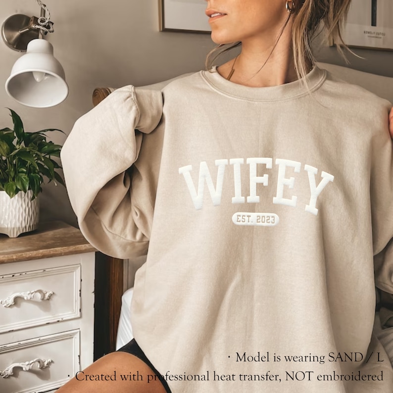 May include: A beige sweatshirt with the text "WIFEY EST. 2023" in white. The sweatshirt is made with a professional heat transfer, not embroidery.