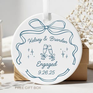 May include: Round white ceramic ornament with a blue ribbon design, the names "Kelsey & Brandon", and the word "Engaged" with the date 9.26.25. Features two champagne glasses and a white ribbon for hanging.