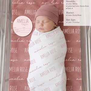 Może przedstawiać: Biały otulacz z nadrukiem imienia Amelia Rose w kolorze brudnego r&oacute;żu, czapeczka w kolorze brudnego r&oacute;żu oraz okrągła tabliczka z imieniem Amelia Rose w kolorze pierwiosnkowym z polami na datę, godzinę, wagę i długość w centymetrach.