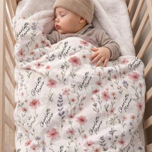 May include: A baby sleeps peacefully in a wooden crib, wrapped in a floral blanket. The blanket features a pattern of pink and purple flowers with the name "Charlotte Marie" printed repeatedly. The baby wears a beige beanie.