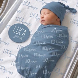 May include: A newborn infant swaddled in a dusty blue blanket featuring the name LUCA James printed in white script. The infant wears a matching blue knotted beanie. A circular blue wooden disc displays the name LUCA James in white text.