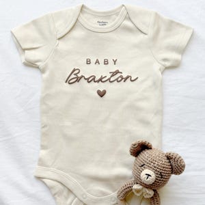 May include: Cream-colored baby onesie with the name "Braxton" in brown script, a brown heart, and the word "BABY". A small, crocheted teddy bear with a bow tie is next to the onesie. The onesie is a size 0-3 months.