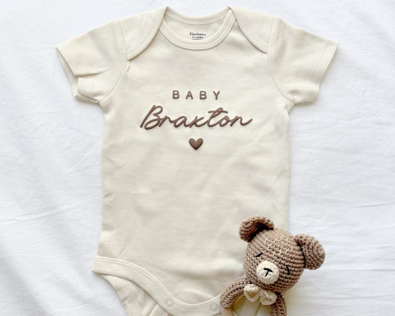 May include: Cream-colored baby onesie with the name "Braxton" in brown script, a brown heart, and the word "BABY". A small, crocheted teddy bear with a bow tie is next to the onesie. The onesie is a size 0-3 months.