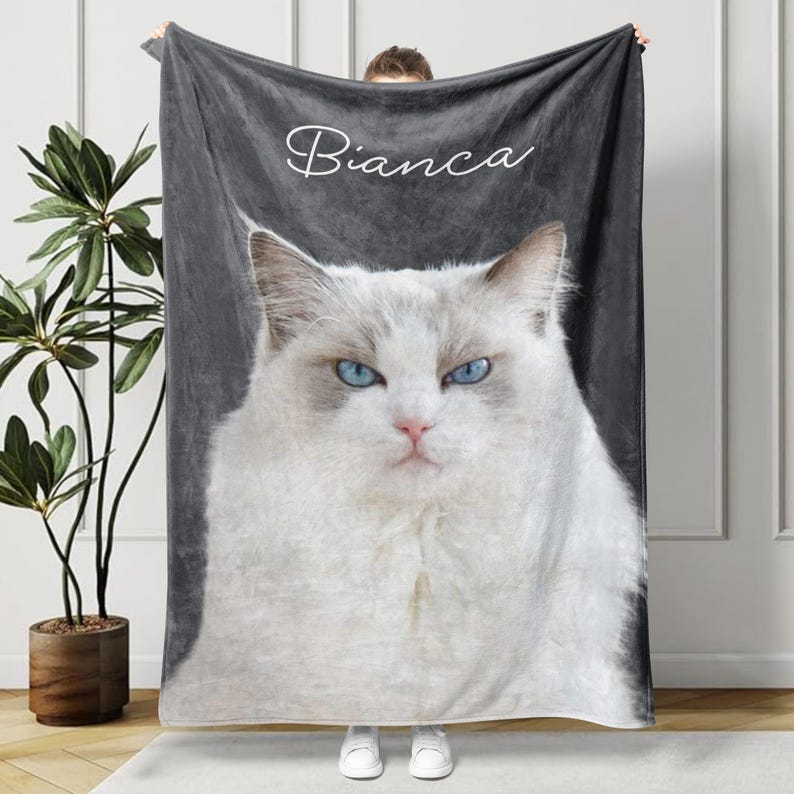 May include: A plush, gray blanket featuring a close-up of a white cat with striking blue eyes and the name "Bianca" in cursive script. The cat has fluffy white fur and a pink nose. The blanket is held up by a person.