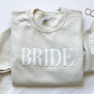 K&ouml;nnte beinhalten: Ein hellbeiger Pullover mit dem Wort "BRIDE" in wei&szlig; auf der Vorderseite gestickt. Der Pullover hat einen Rundhalsausschnitt und lange &Auml;rmel. Der &Auml;rmel hat ein kleines Herz mit den Buchstaben "T" und "M" gestickt.