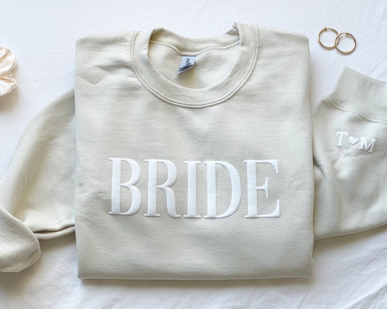 K&ouml;nnte beinhalten: Ein hellbeiger Pullover mit dem Wort "BRIDE" in wei&szlig; auf der Vorderseite gestickt. Der Pullover hat einen Rundhalsausschnitt und lange &Auml;rmel. Der &Auml;rmel hat ein kleines Herz mit den Buchstaben "T" und "M" gestickt.