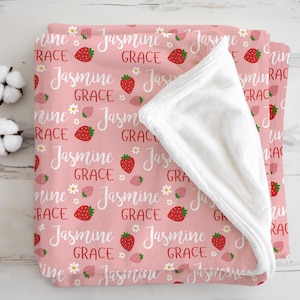 Könnte beinhalten: Eine rosa Babydecke mit einem Muster aus Erdbeeren, Gänseblümchen und dem Namen "Jasmine Grace" in weißer Schrift. Die Decke hat eine weiche, weiße, plüschige Unterseite.
