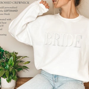 K&ouml;nnte beinhalten: Wei&szlig;er Crewneck-Pullover mit dem Wort "BRIDE" auf der Vorderseite. Der Pullover wird von einem Model getragen.