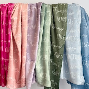 May include: A display of personalized blankets in various colors, including pink, peach, lavender, green, olive, light blue, and blue. Each blanket is printed with a different name in white script. The blankets are soft and plush.