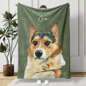 May include: A green blanket featuring a watercolor portrait of a Corgi dog. The dog has brown, white, and black fur. The name "Leo" is written in cursive at the top of the blanket. The blanket is held up by a person.