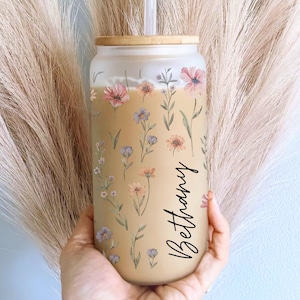 K&ouml;nnte beinhalten: Ein Glasbecher mit Bambusdeckel und Strohhalm, verziert mit einem floralen Muster in Pastellt&ouml;nen. Auf dem Becher steht der Name "Bethany" in einer Schreibschrift. Das Design umfasst rosa, lila und gelbe Blumen mit gr&uuml;nen Bl&auml;ttern.