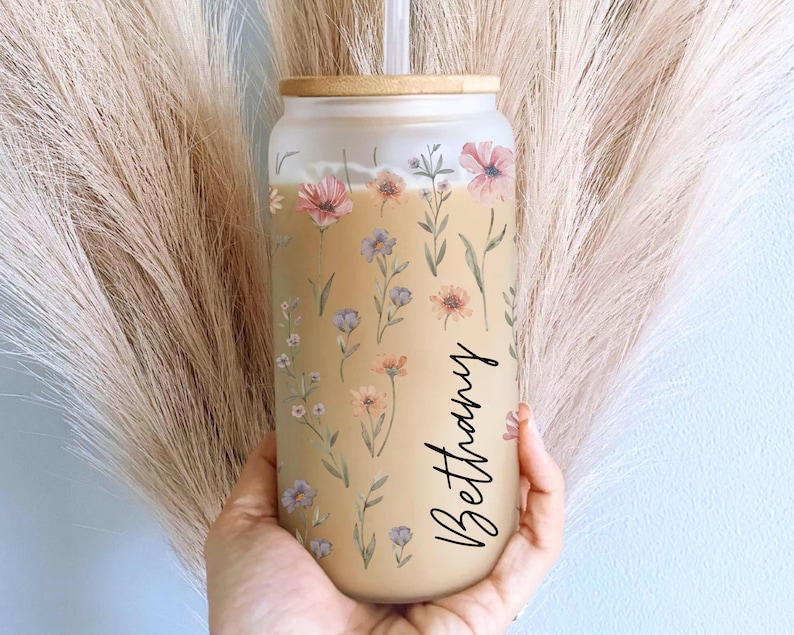 K&ouml;nnte beinhalten: Ein Glasbecher mit Bambusdeckel und Strohhalm, verziert mit einem floralen Muster in Pastellt&ouml;nen. Auf dem Becher steht der Name "Bethany" in einer Schreibschrift. Das Design umfasst rosa, lila und gelbe Blumen mit gr&uuml;nen Bl&auml;ttern.