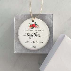 Puede incluir: Un adorno navide&ntilde;o de cer&aacute;mica blanca con un dise&ntilde;o floral y el texto "OUR FIRST CHRISTMAS together DAVID & ANNA -2021-". El adorno es redondo y viene en una caja de regalo blanca con una cinta dorada.