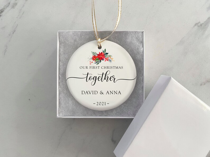Puede incluir: Un adorno navide&ntilde;o de cer&aacute;mica blanca con un dise&ntilde;o floral y el texto "OUR FIRST CHRISTMAS together DAVID & ANNA -2021-". El adorno es redondo y viene en una caja de regalo blanca con una cinta dorada.