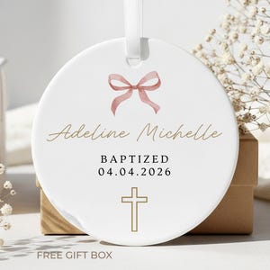 Peut inclure: Ornement de bapt&ecirc;me en c&eacute;ramique blanche avec un n&oelig;ud rose, le nom "Adeline Michelle", le mot "BAPTIZED", la date "04.04.2026" et une croix dor&eacute;e. L'ornement a un ruban blanc pour l'accrocher. Le texte "FREE GIFT BOX" est en bas.