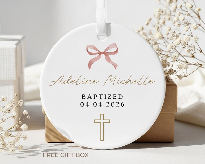 Peut inclure: Ornement de bapt&ecirc;me en c&eacute;ramique blanche avec un n&oelig;ud rose, le nom "Adeline Michelle", le mot "BAPTIZED", la date "04.04.2026" et une croix dor&eacute;e. L'ornement a un ruban blanc pour l'accrocher. Le texte "FREE GIFT BOX" est en bas.