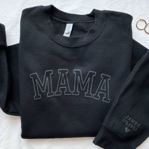 May include: Black sweatshirt with the word "MAMA" in large, white, outlined letters. The sleeve has the names "JAMES" and "OLIVIA" with a heart symbol.