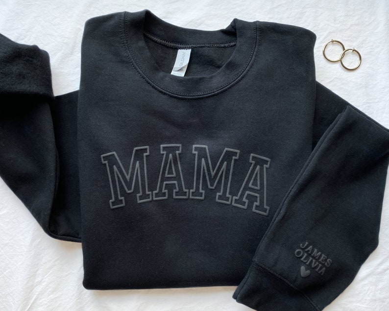 May include: Black sweatshirt with the word "MAMA" in large, white, outlined letters. The sleeve has the names "JAMES" and "OLIVIA" with a heart symbol.