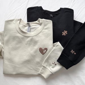 May include: Three sweatshirts in cream and black, with embroidered details. The cream sweatshirt has a heart with a puzzle piece, the black one has a puzzle piece and the initial "M" on the sleeve. The other sleeve has the initials "J" and a heart.